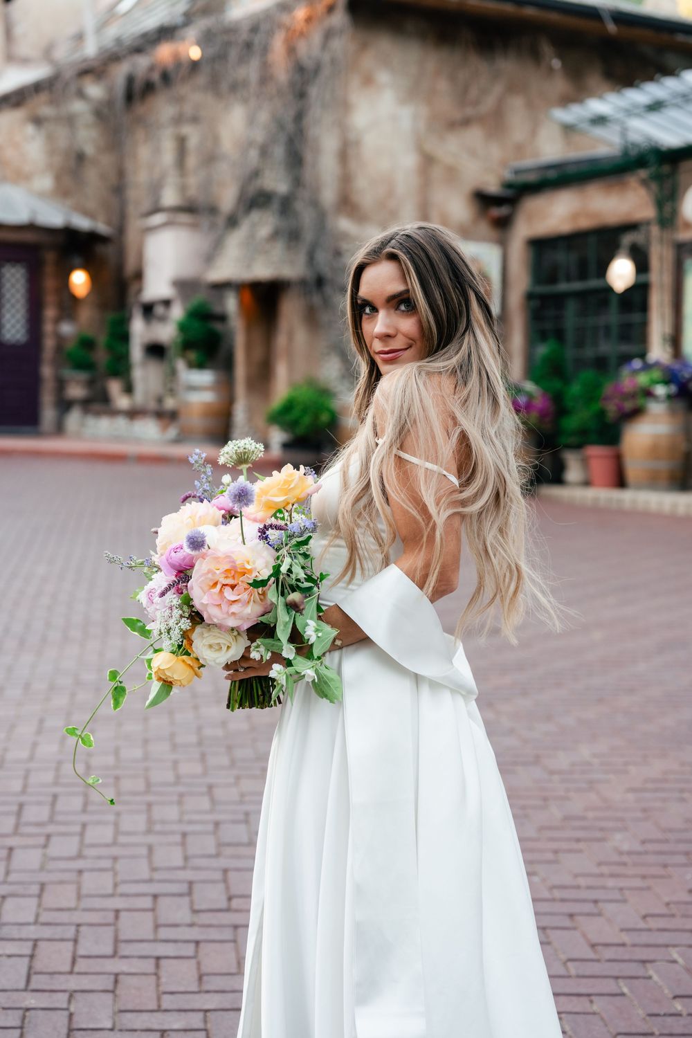 Woman in flowing white dress holds colorful bouquet against rustic building backdrop.