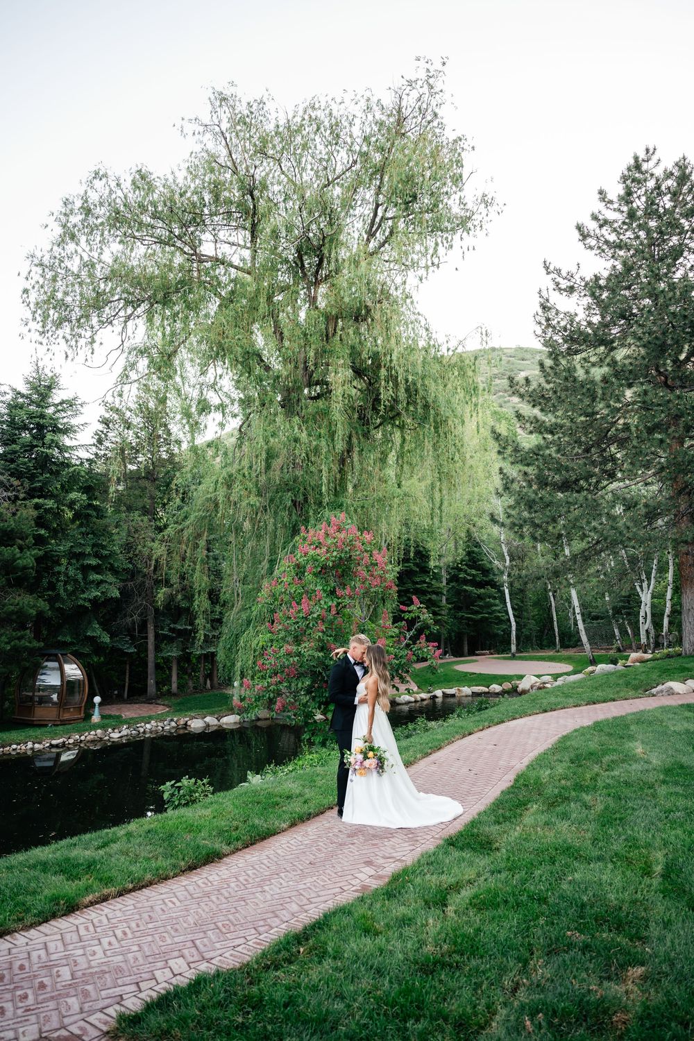 A couple embraces on a brick path surrounded by lush gardens with a dramatic weeping willow tree overhead.