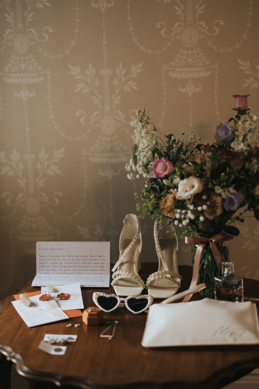 Wedding details arranged on wooden surface including retro sunglasses, floral arrangements, invitations and elegant bridal shoes.