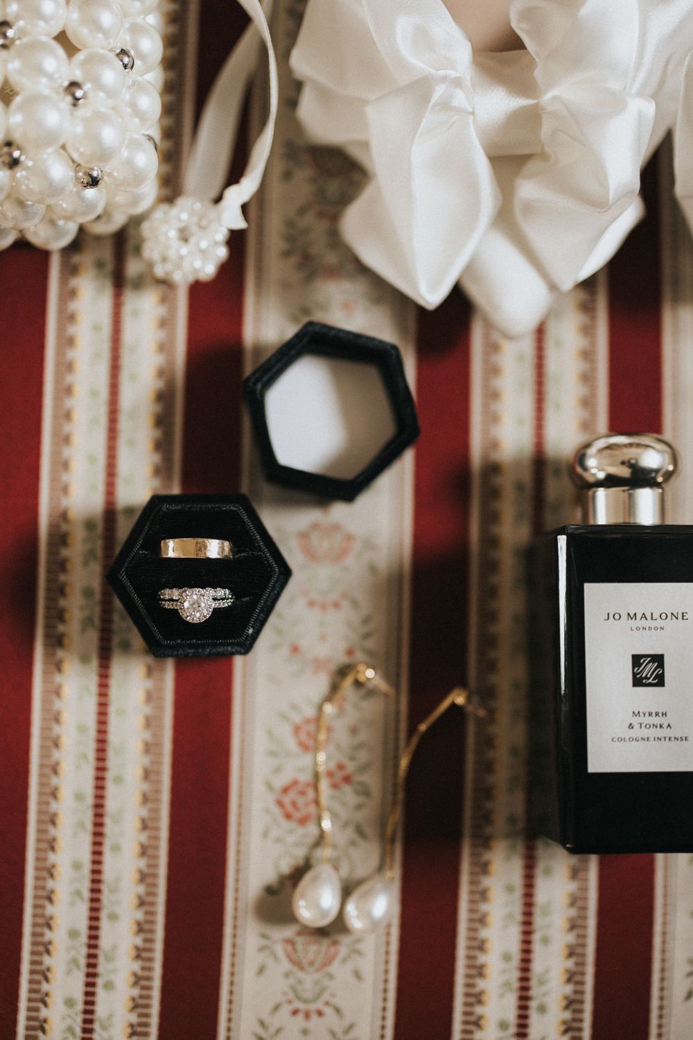 Wedding rings in black hexagonal box with Jo Malone perfume bottle and white floral accents on striped fabric backdrop.