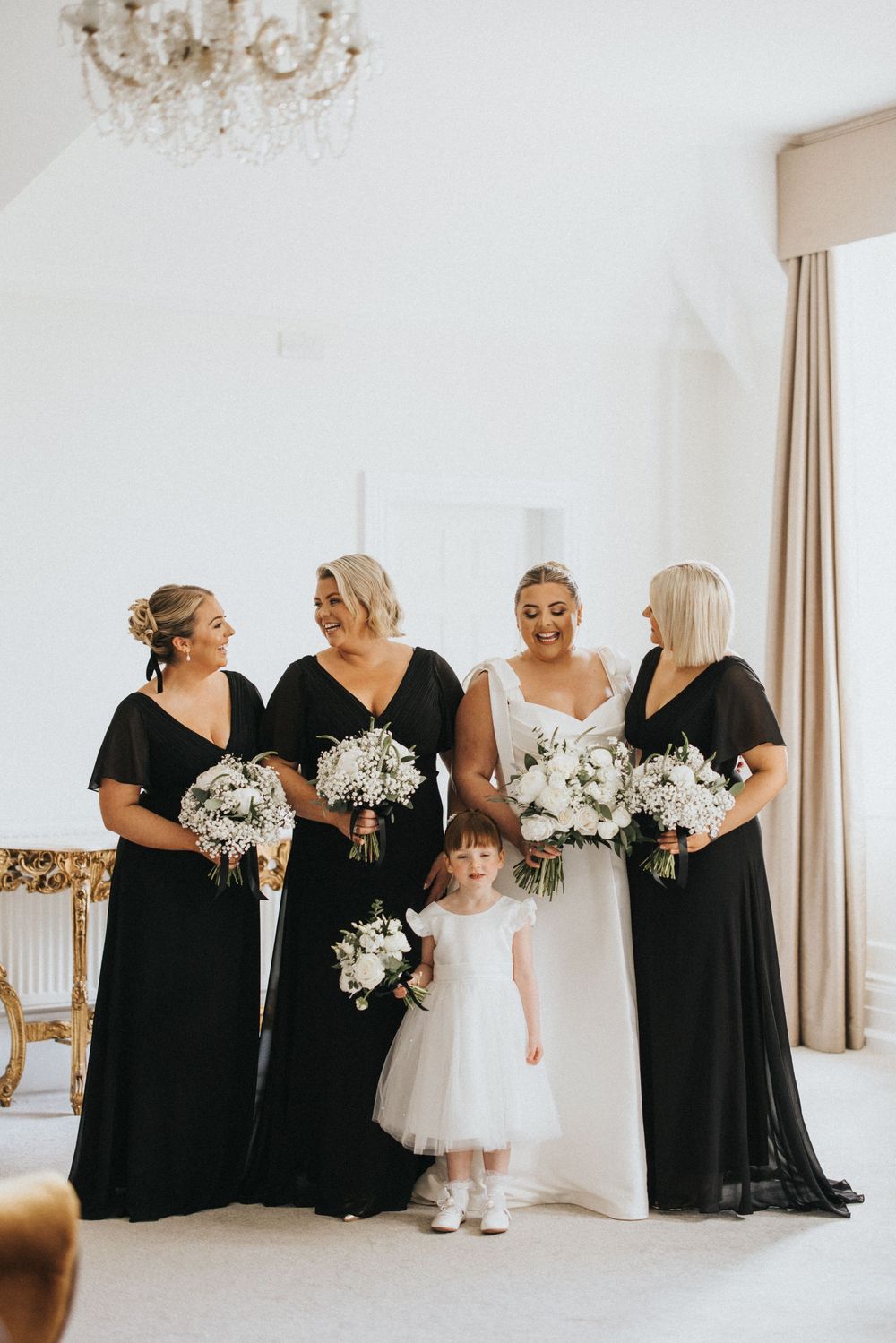 Wedding party in black dresses and white bouquets pose with flower girl in tulle dress during elegant indoor ceremony.