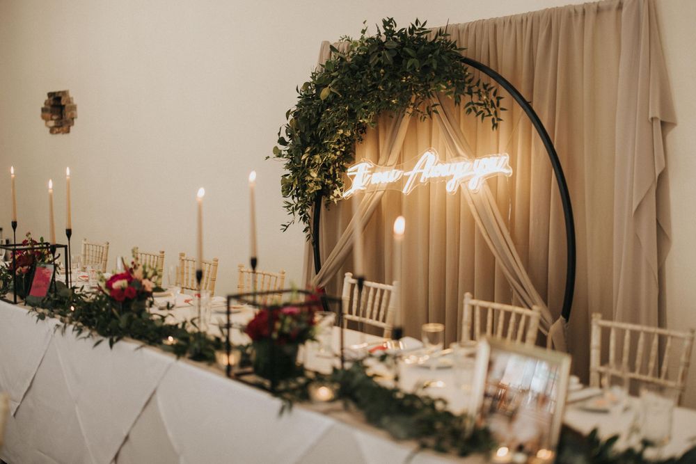 Wedding reception table setting with arch decorated with greenery, neon sign and white candles along elegant tablescape.
