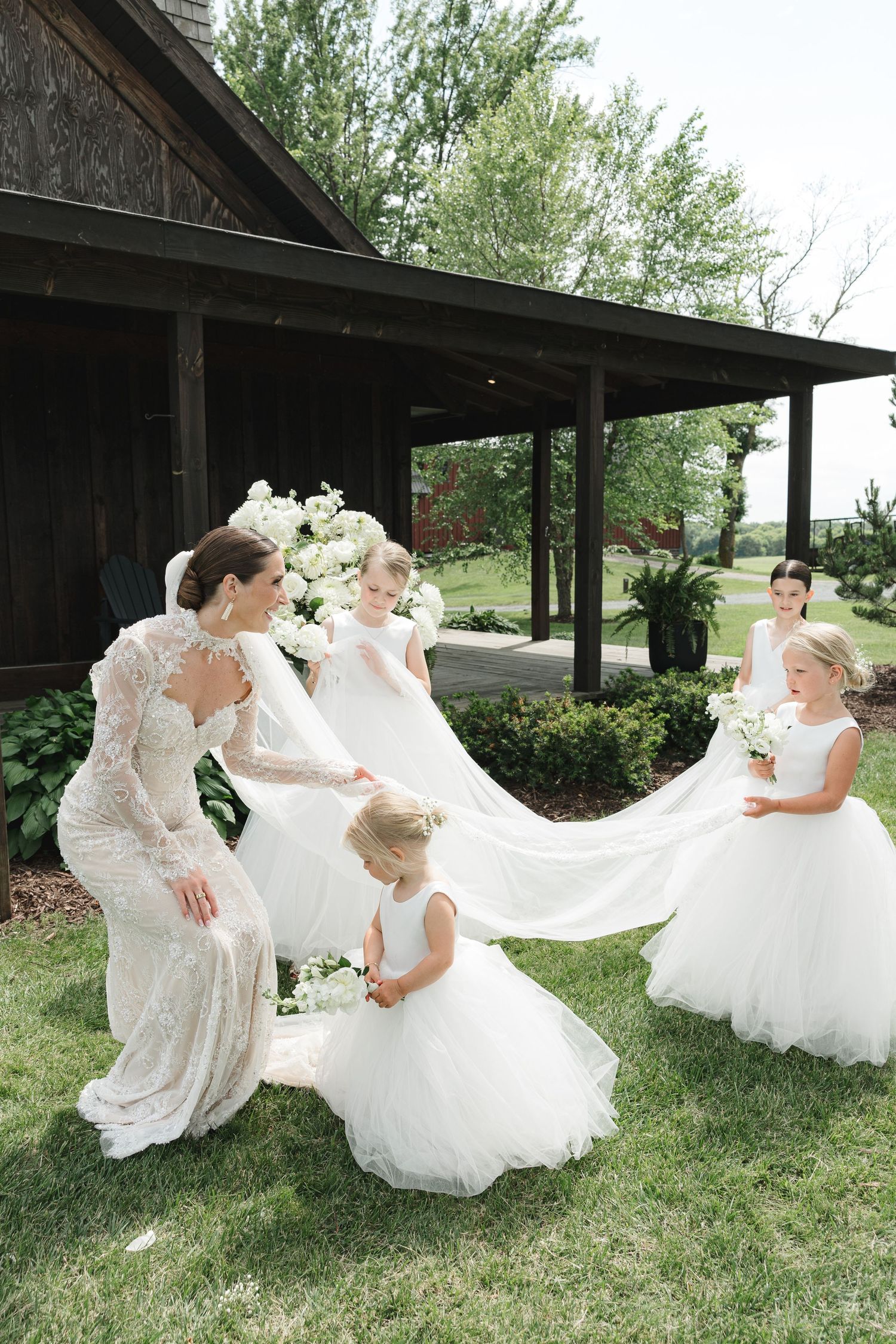 A group of bridesmaids in white dresses help adjust a bride's flowing veil outside a rustic cabin on a grassy lawn.