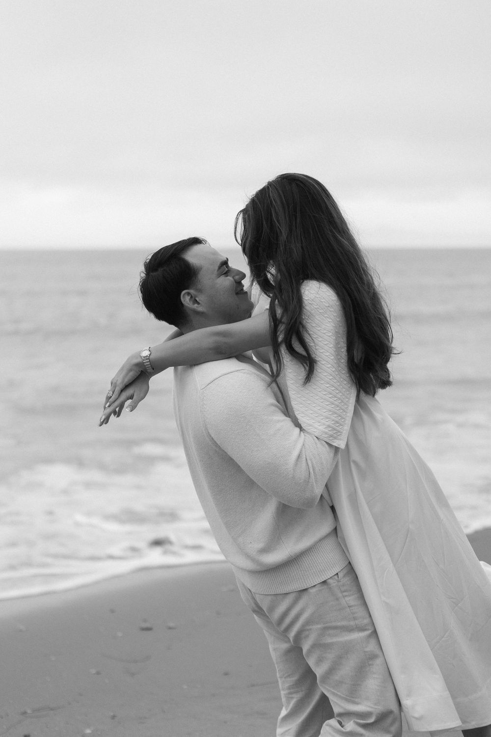 Backlit photo of couple hugging with ocean and clouds in background.