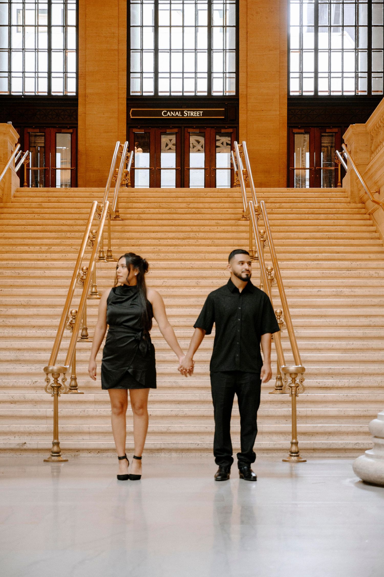 A couple in elegant dark attire stands holding hands on grand marble stairs with brass railings inside a historic building.