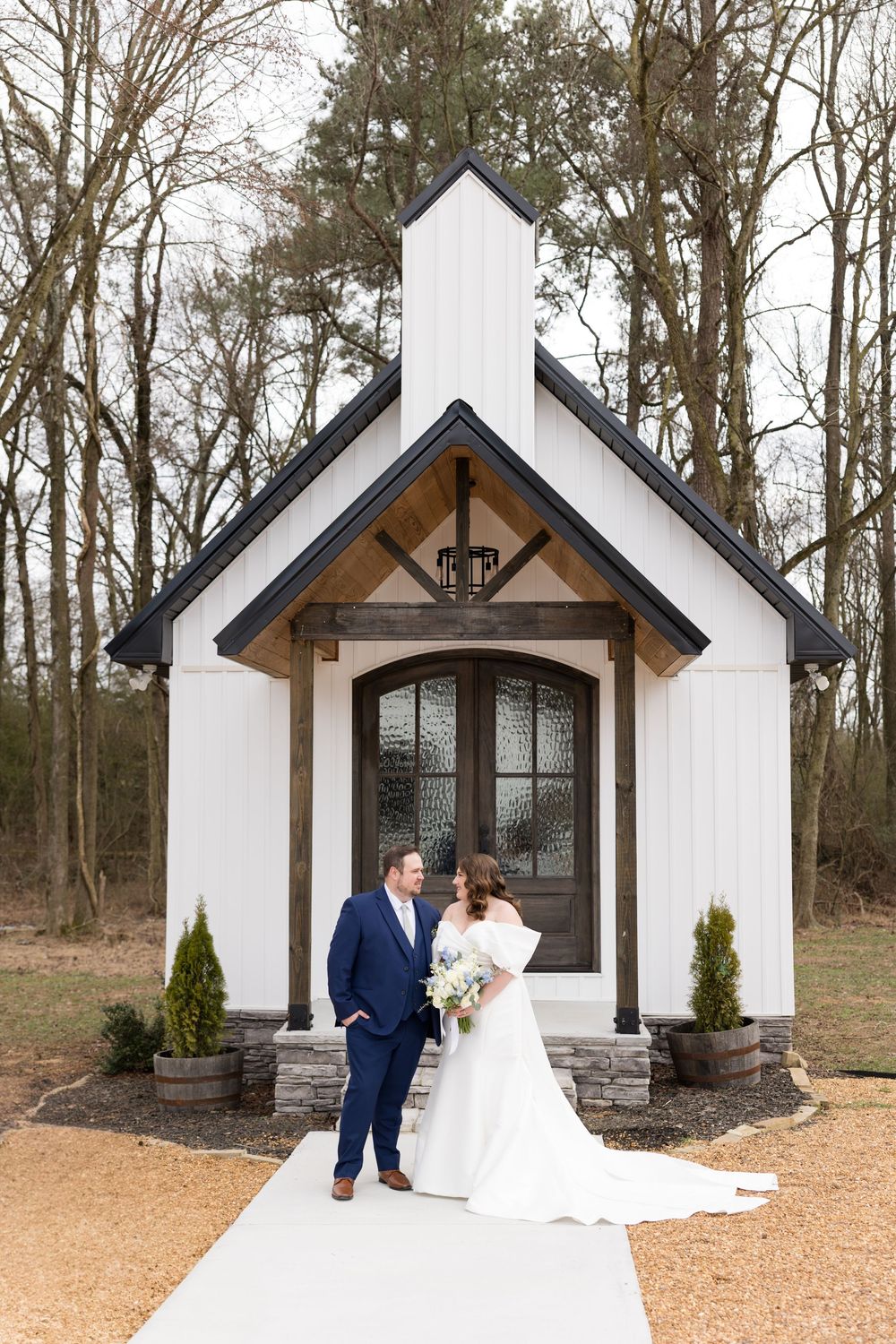 Four white chapel buildings with timber accents and dark windows stand in a wooded setting at sunset.