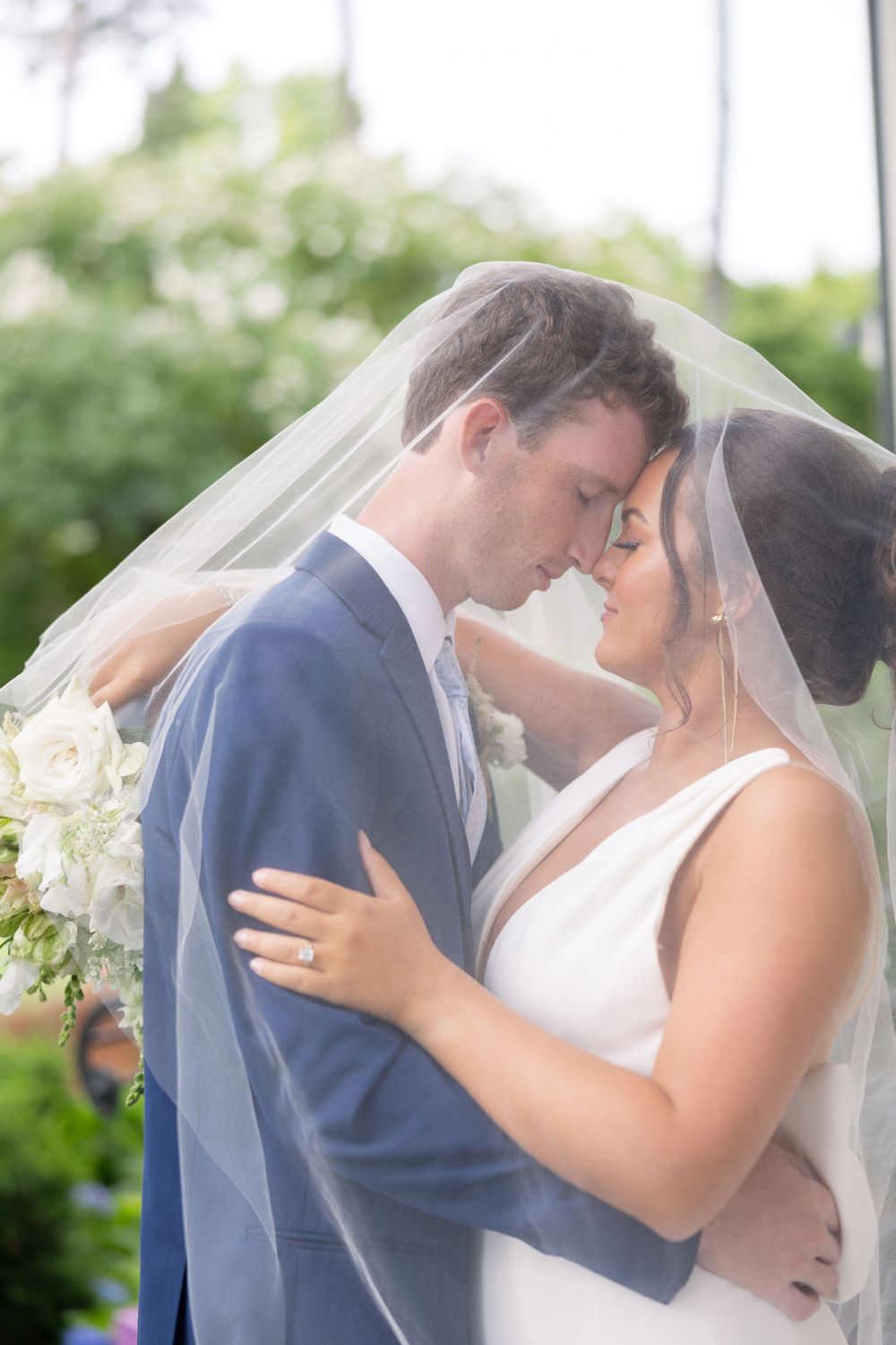 Romantic wedding couple embrace under flowing veil in outdoor garden setting at sunset.