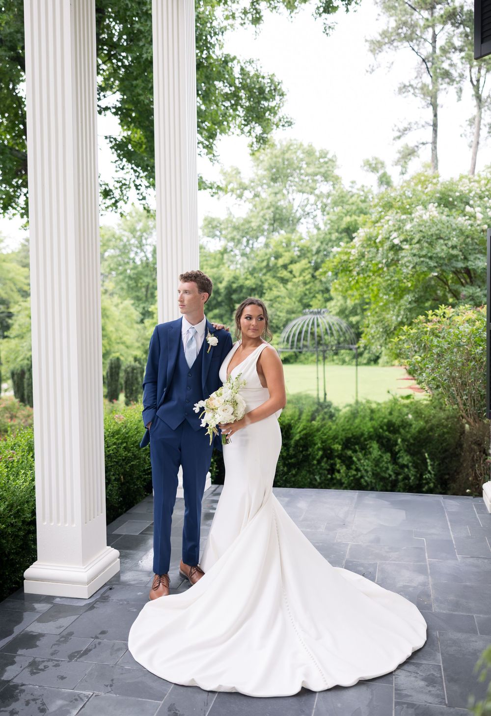 Wedding couple poses on a stone patio with elegant white columns and lush greenery in the background.