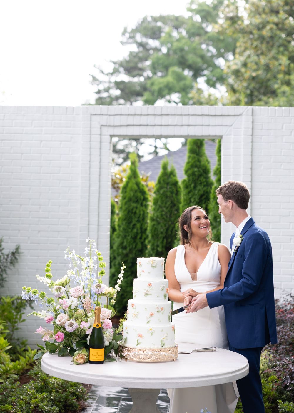 A couple stands together at their outdoor wedding reception beside an elegant white tiered cake and champagne bottle.