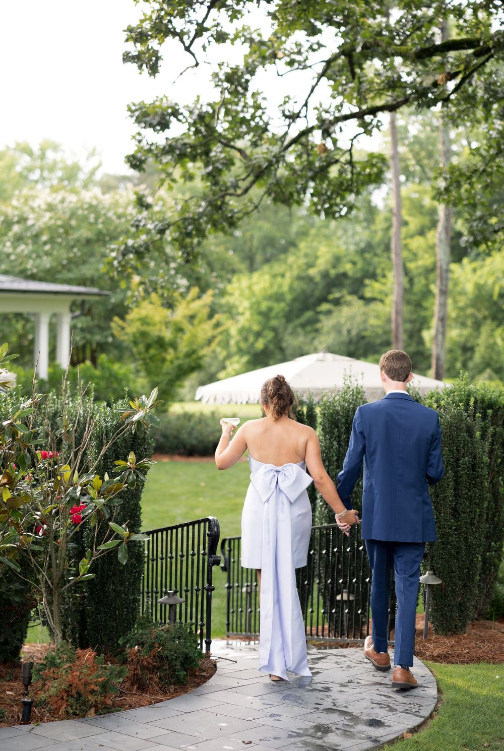 A couple in wedding attire walks along a stone path beside manicured hedges and a white gazebo.