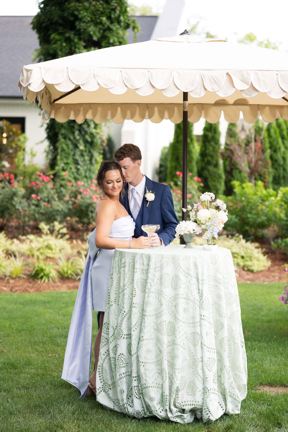 A couple stands under an elegant cream-colored umbrella by a decorative table at an outdoor garden wedding reception.