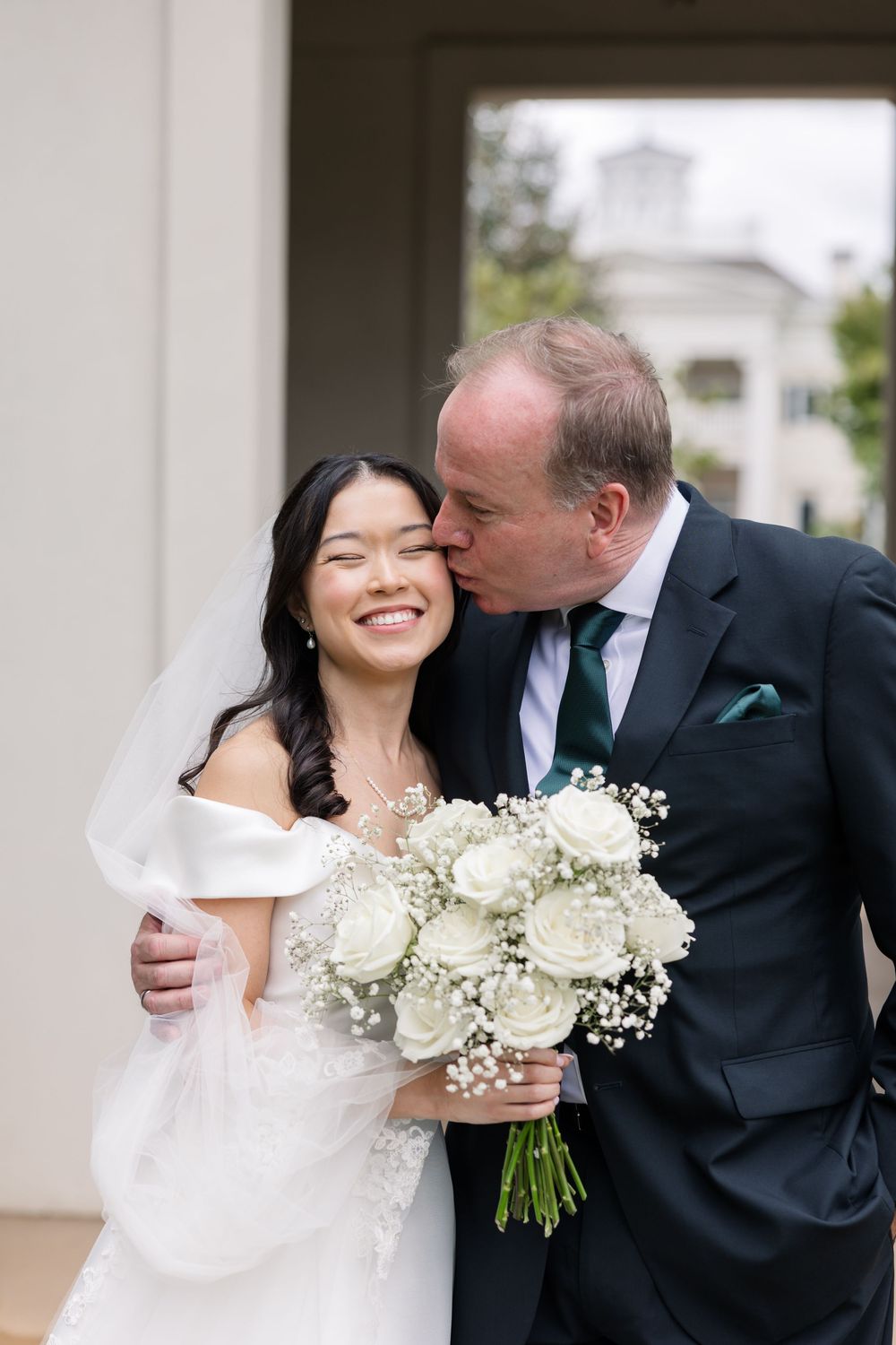 A joyful wedding couple embraces outdoors while holding a beautiful white rose and baby's breath bouquet.