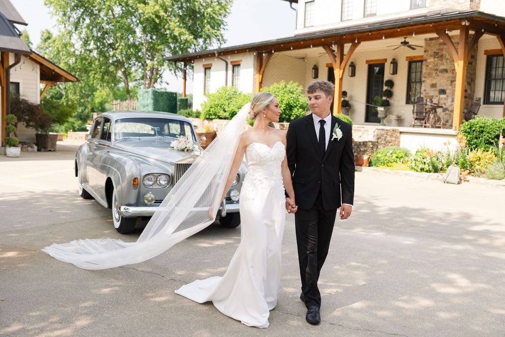 A joyful newlywed couple walks together on a sunny day with a classic vintage car and stone building in the background.