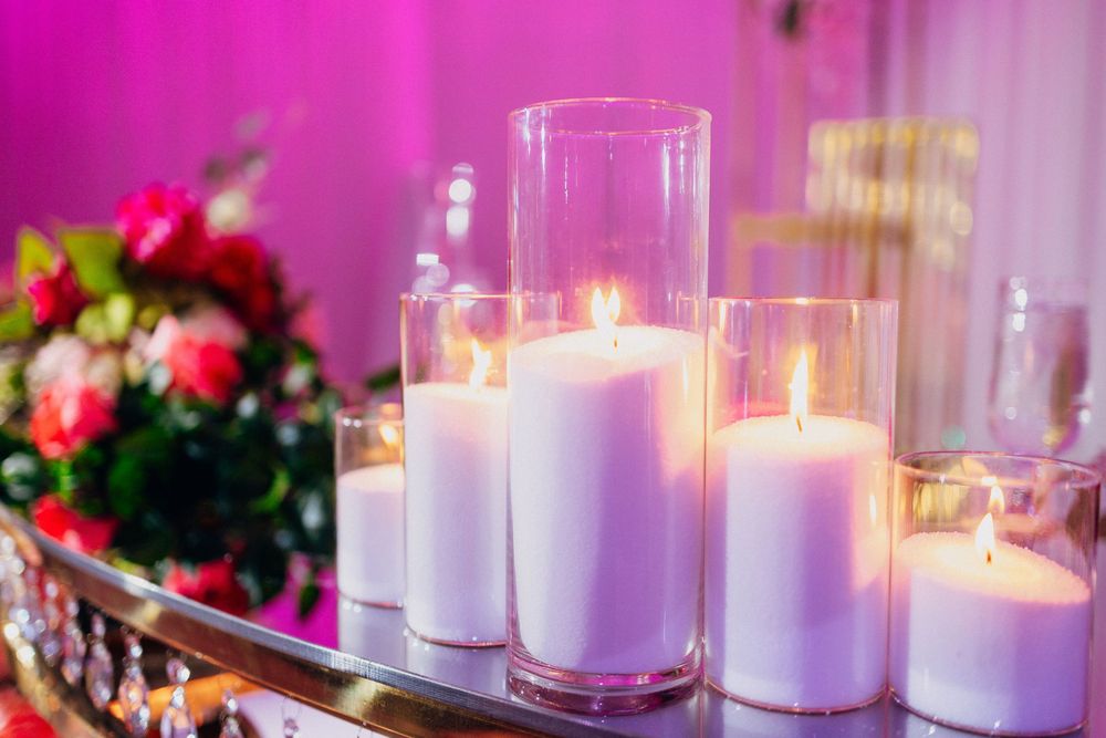 Elegant white pillar candles in glass cylinders arranged on mirrored surface with pink floral decor in background.