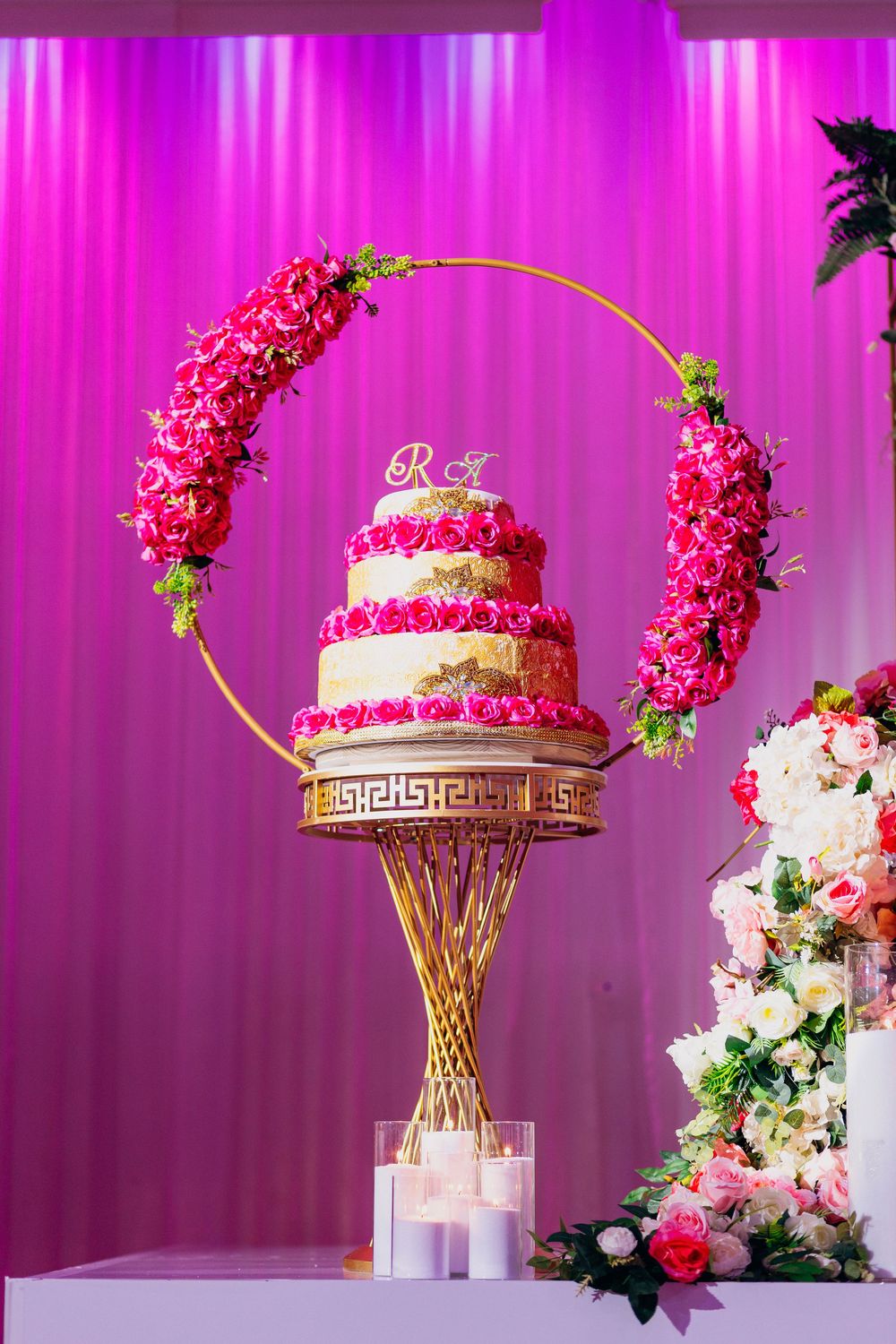 Elegant wedding cake with pink floral decorations displayed on gold stand against magenta backdrop with cascading flowers.