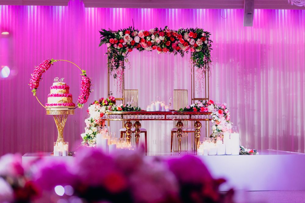 A luxurious pink-lit wedding reception venue with floral decorations, white drapery, and an elegant cake display.