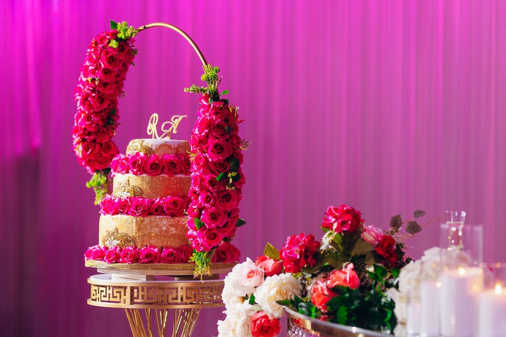 Pink and red floral decorations adorn an elegant wedding cake with gold accents against a vibrant purple backdrop.