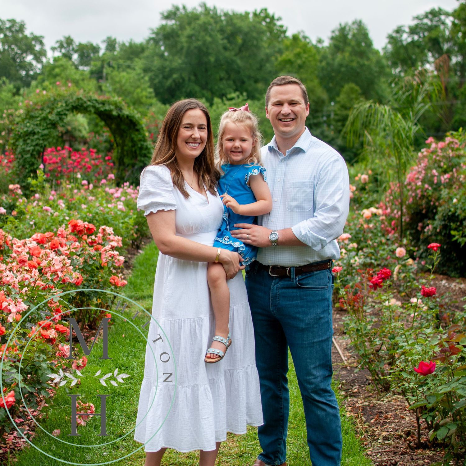 A family poses together in a blooming rose garden with vibrant red and pink flowers on a summer day.