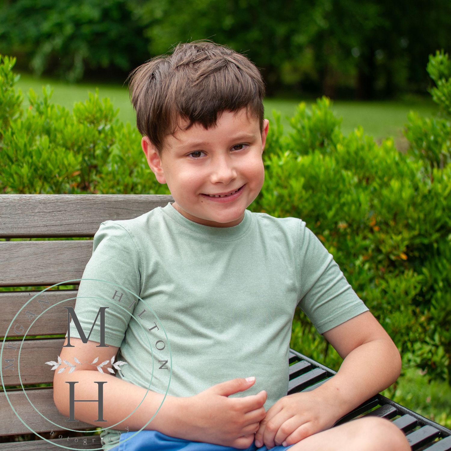 Young child in a mint green t-shirt sits relaxed on a wooden bench with green bushes and trees in the background.