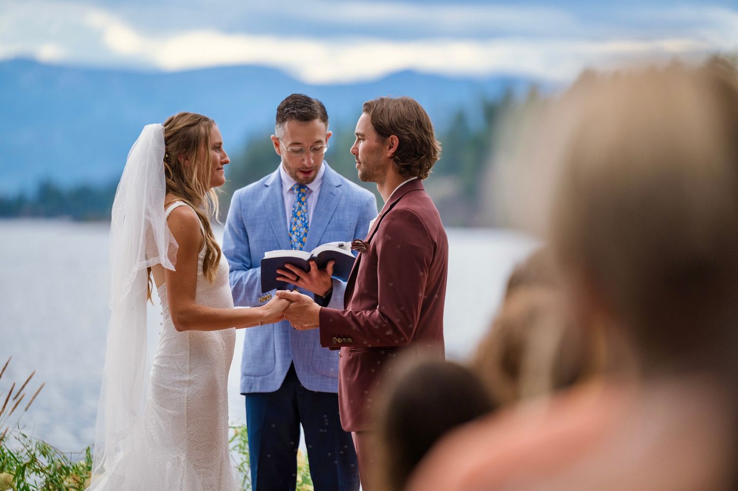 A couple holds hands during an outdoor wedding ceremony beside a serene lake with mountains in the background.