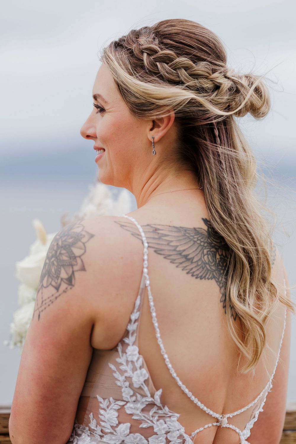 Elegant bridal hairstyle featuring a braided crown and loose waves with visible wing tattoo and lace dress detail.