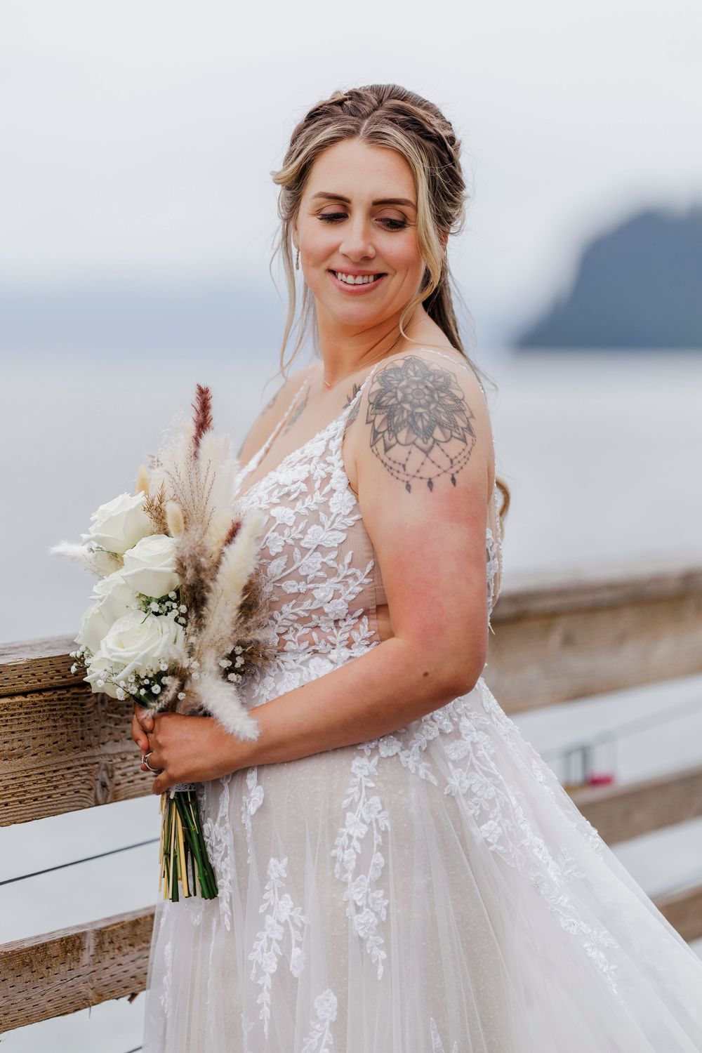 A bride in a lace wedding dress with a shoulder tattoo holds a bouquet of white flowers and pampas grass by the water.