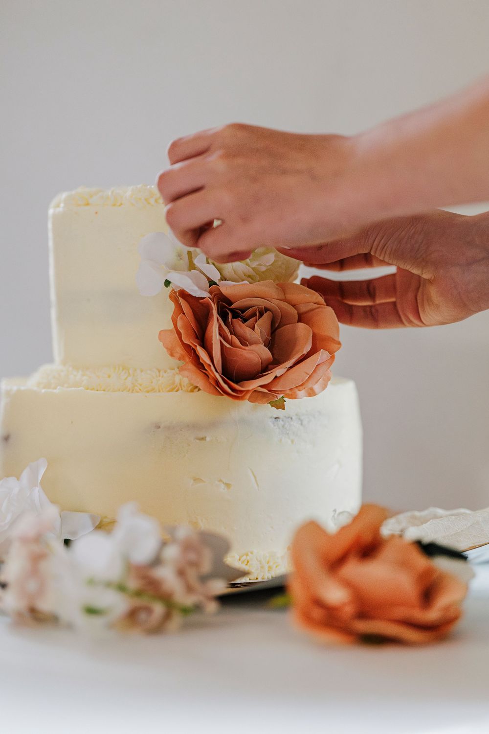 Hands decorating a white tiered wedding cake with peach-colored roses.