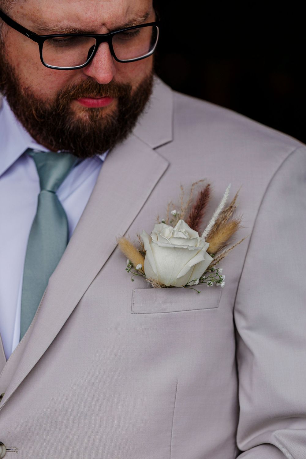 Close-up of a white rose boutonniere with dried wheat accents on a light gray suit lapel.