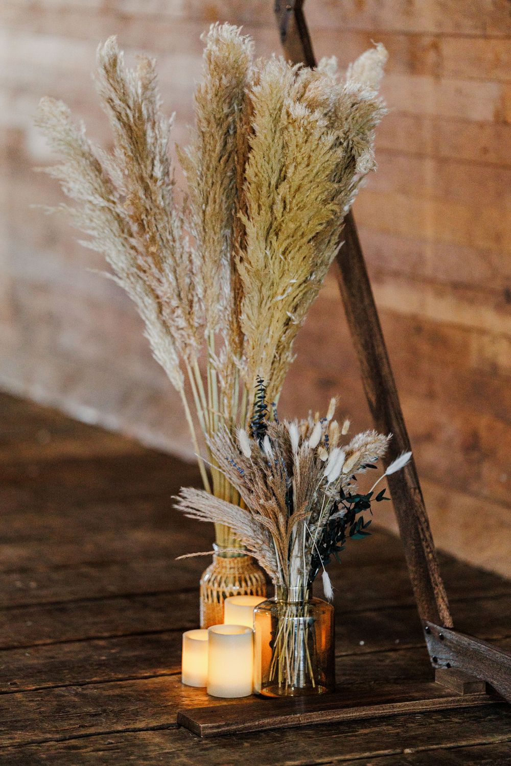 Dried wheat and pampas grass arrangements in glass vases with glowing candles create a rustic wedding decor setting.
