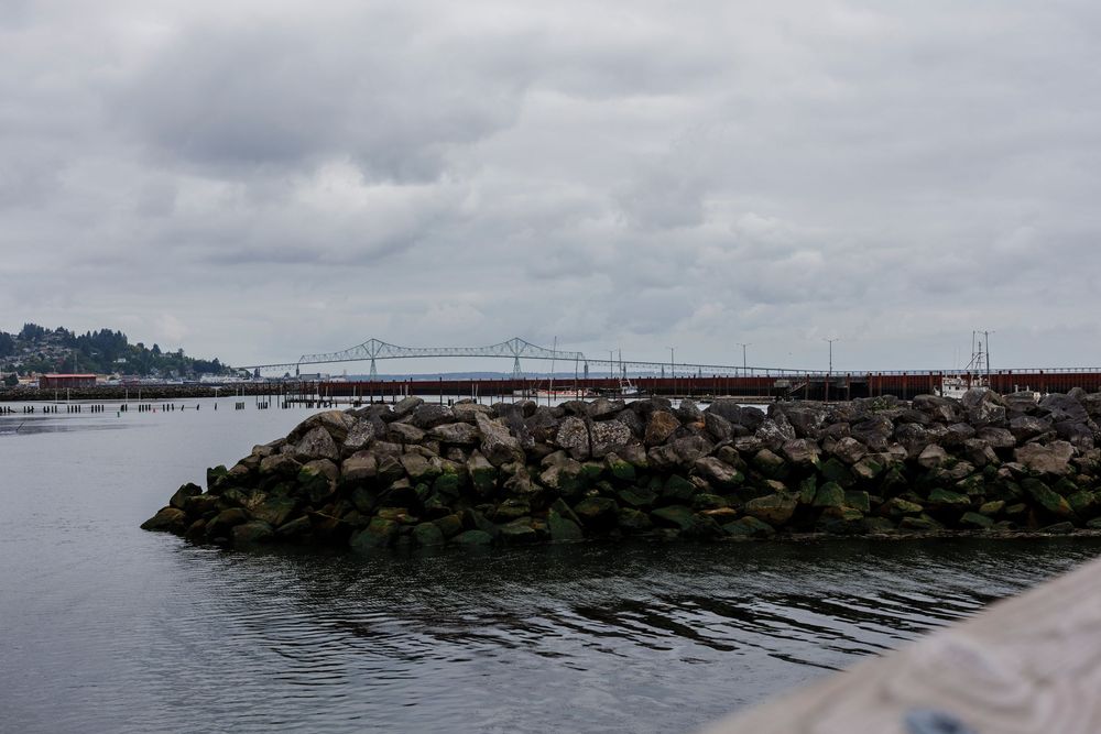 Rocky breakwater stretches into a misty gray ocean with boats and buildings visible on the distant shoreline.