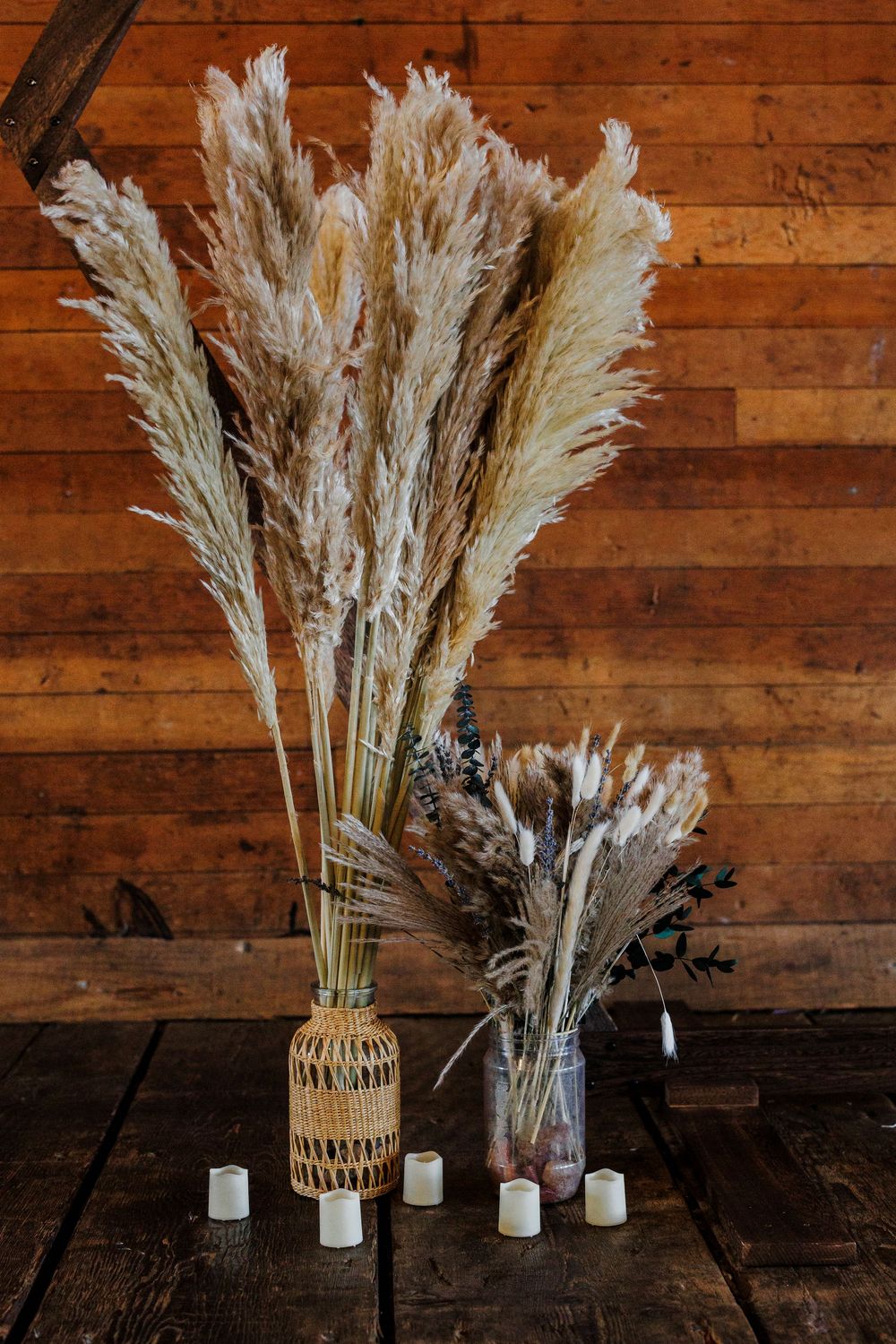 Dried pampas grass and floral arrangements in woven baskets against rustic wooden wall with candles.