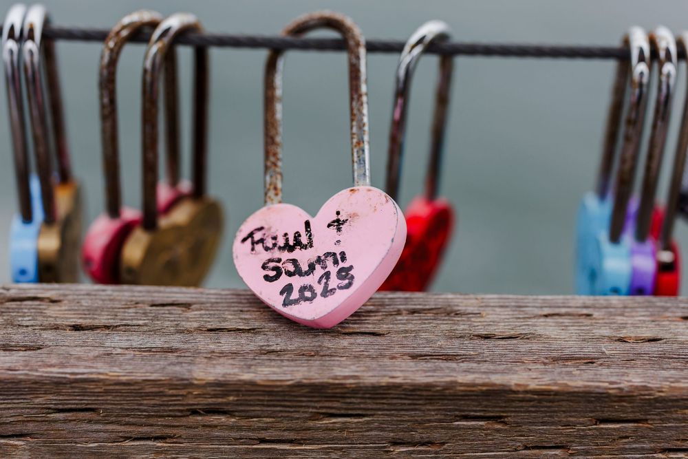 A pink heart-shaped love lock with text inscribed on weathered wood.