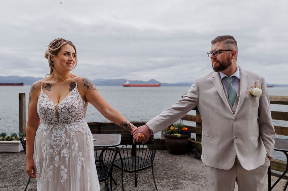 Wedding couple holding hands on a waterfront deck with ships visible in the distance.