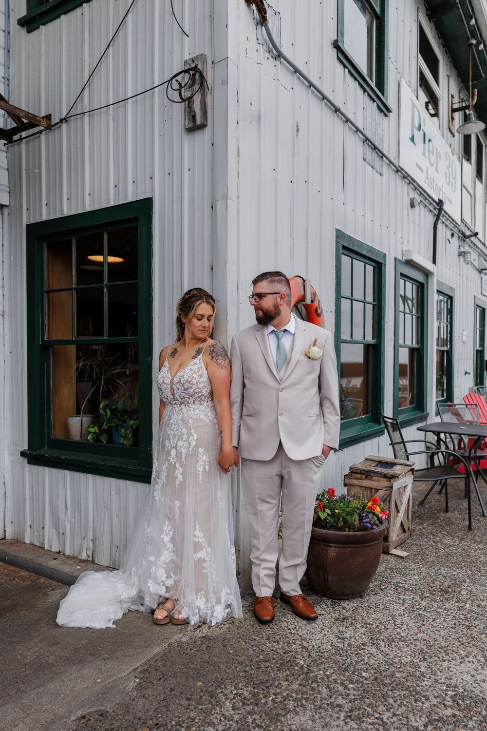 Newlyweds pose together outside a white industrial building with green trim and potted flowers.