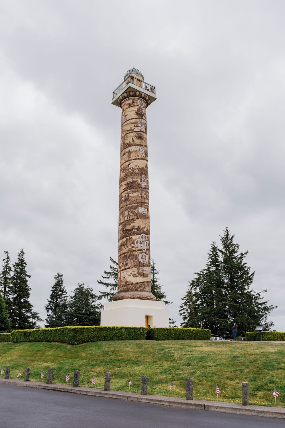 Tall decorative stone column monument surrounded by evergreen trees under cloudy skies.