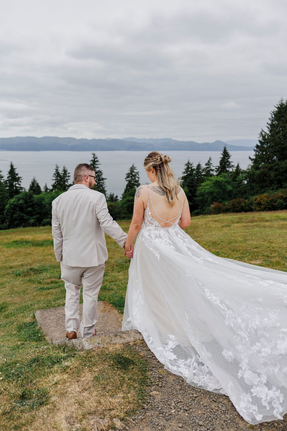 A wedding couple walks along a hilltop path with a flowing white lace dress trailing behind them.