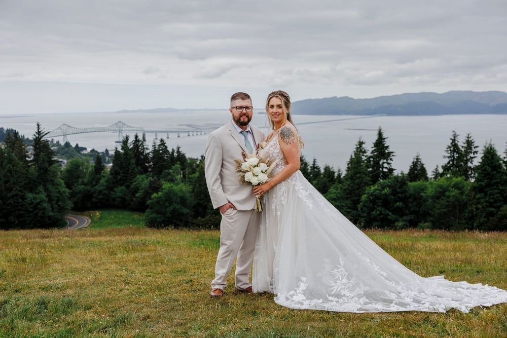 A couple in wedding attire stands on a hilltop overlooking the bay with evergreen trees in the background.