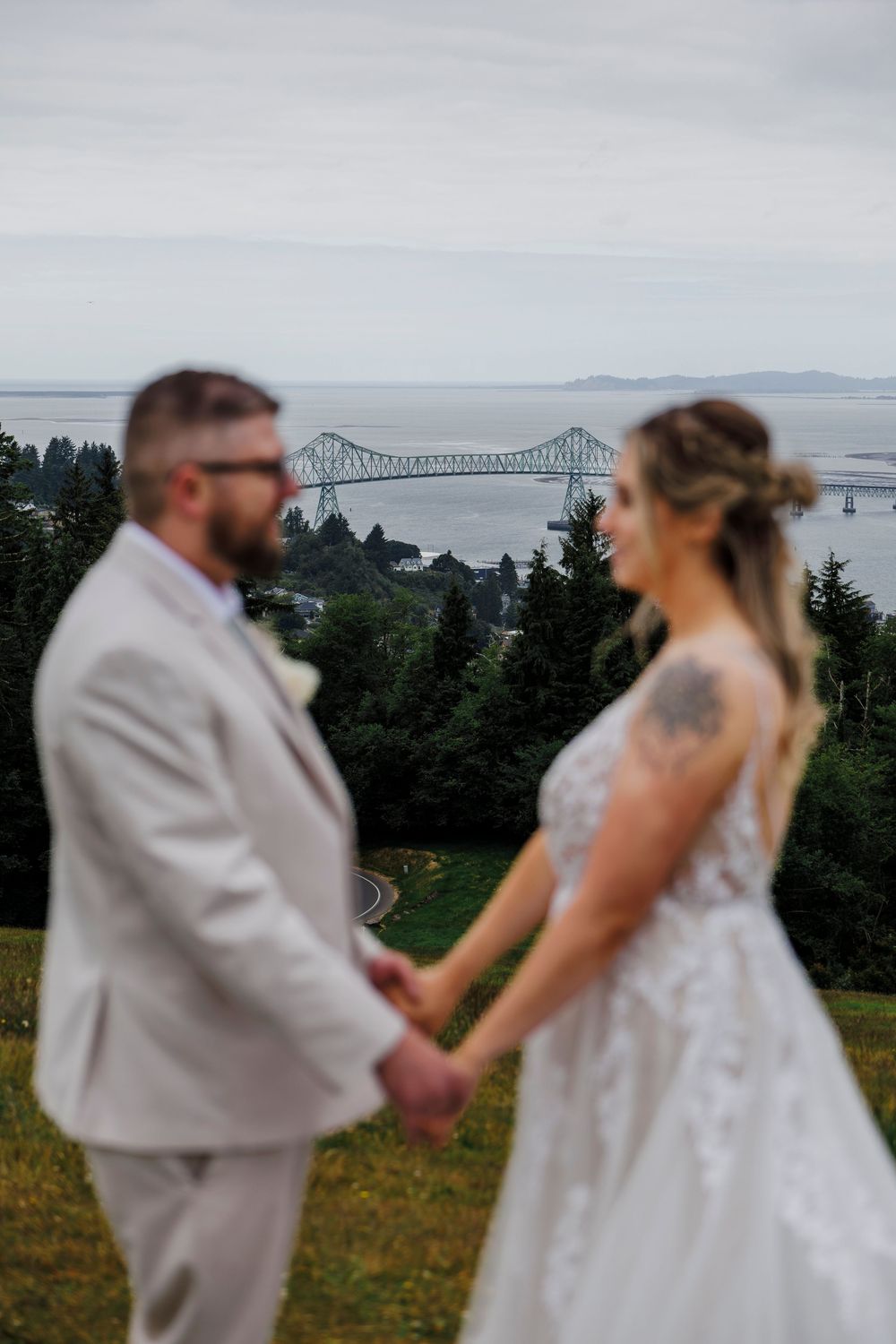 Romantic wedding portrait with Astoria-Megler Bridge visible in background through trees.