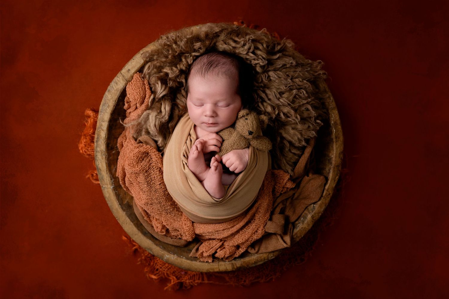 Sleeping newborn baby wrapped in beige fabric nestled in round wooden bowl against red background.