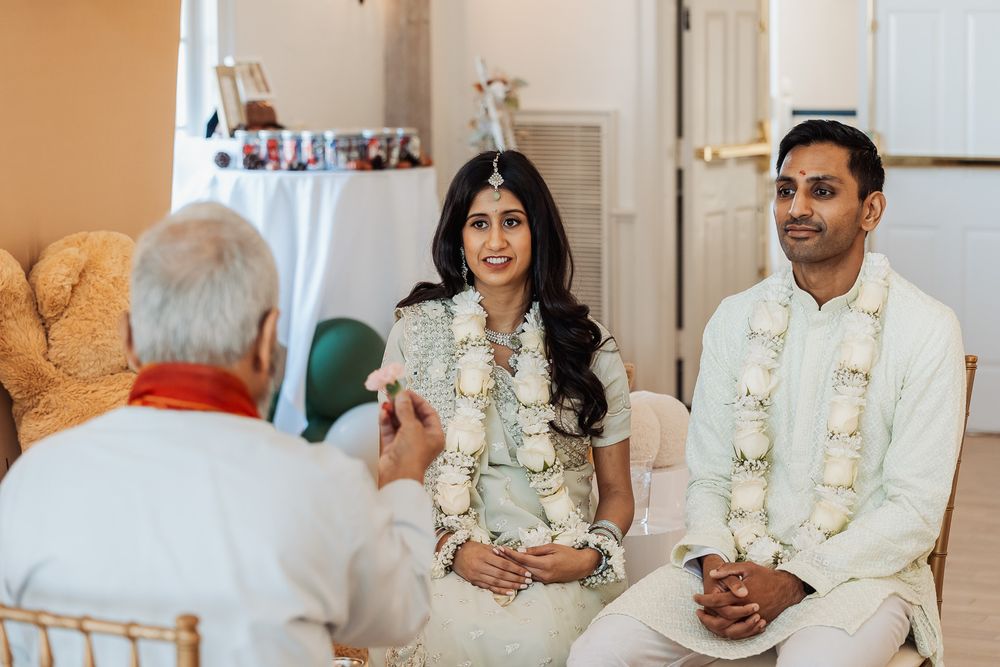 A couple in traditional white wedding attire with floral garlands participates in an Indian ceremony with an officiant.