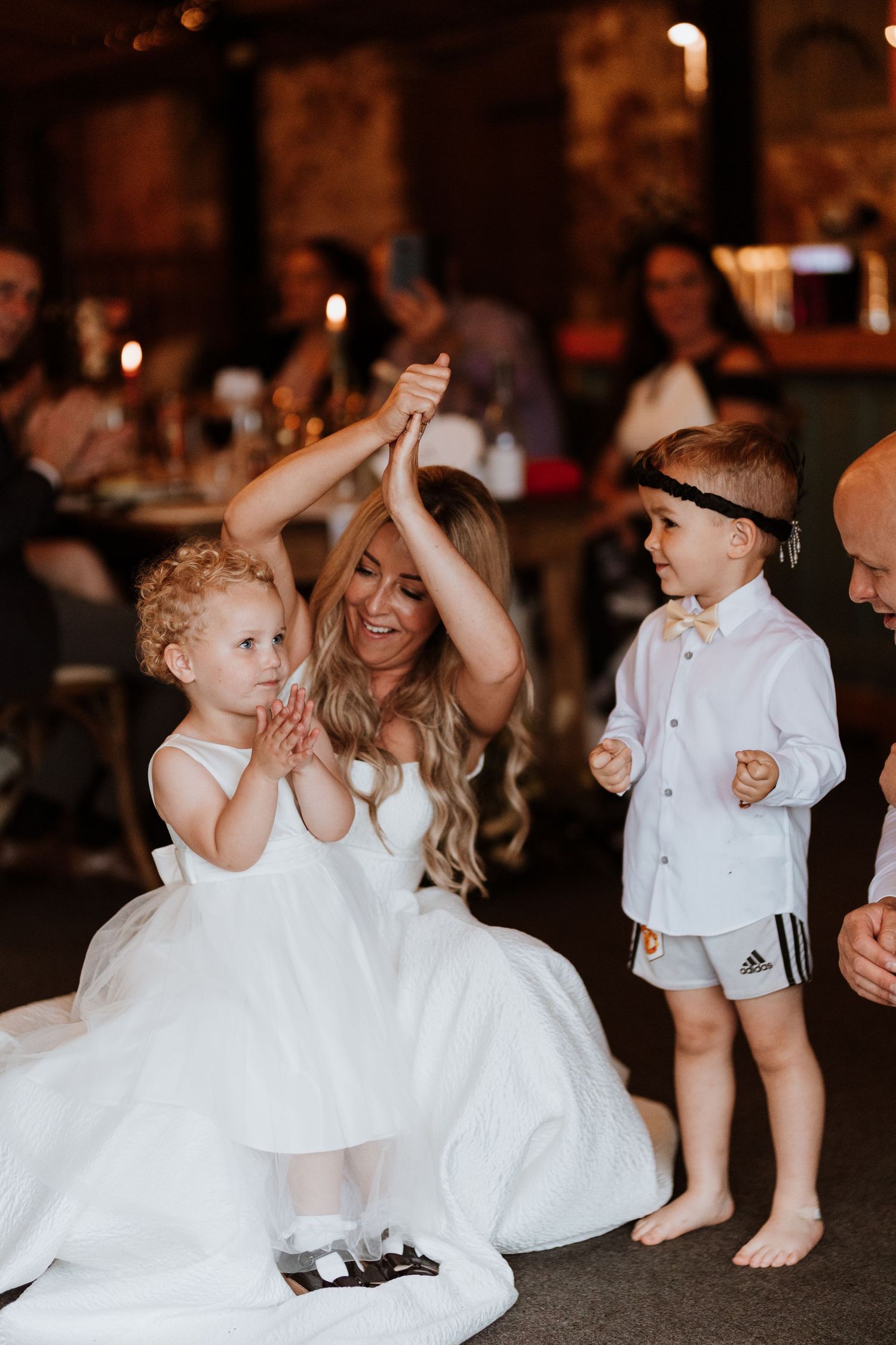 A person in a white wedding dress dances with a young child while another small child watches at a reception.