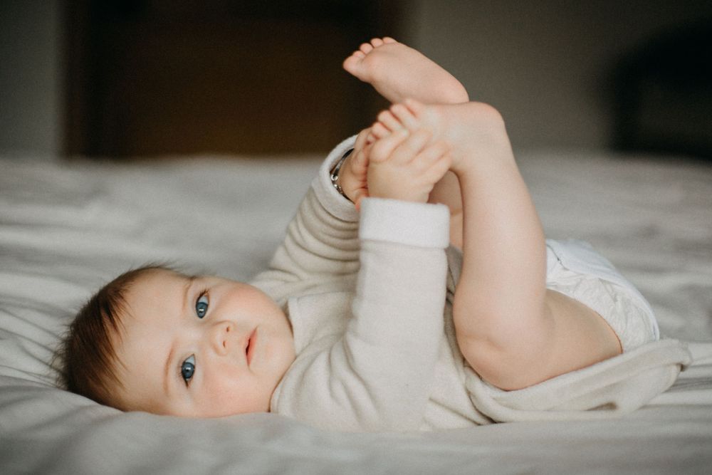 Baby in white onesie playing with feet while lying on bed.