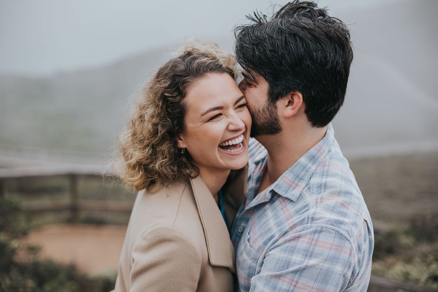 Candid engagement photo of a couple laughing in a dreamy mountain landscape during golden hour, capturing love and natural beauty.