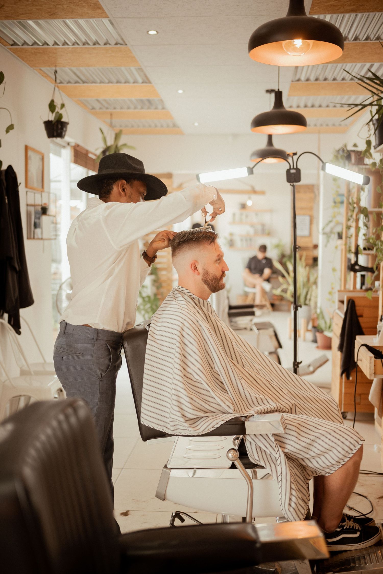 Barber in wide-brimmed hat cuts client's hair in modern, plant-decorated salon.