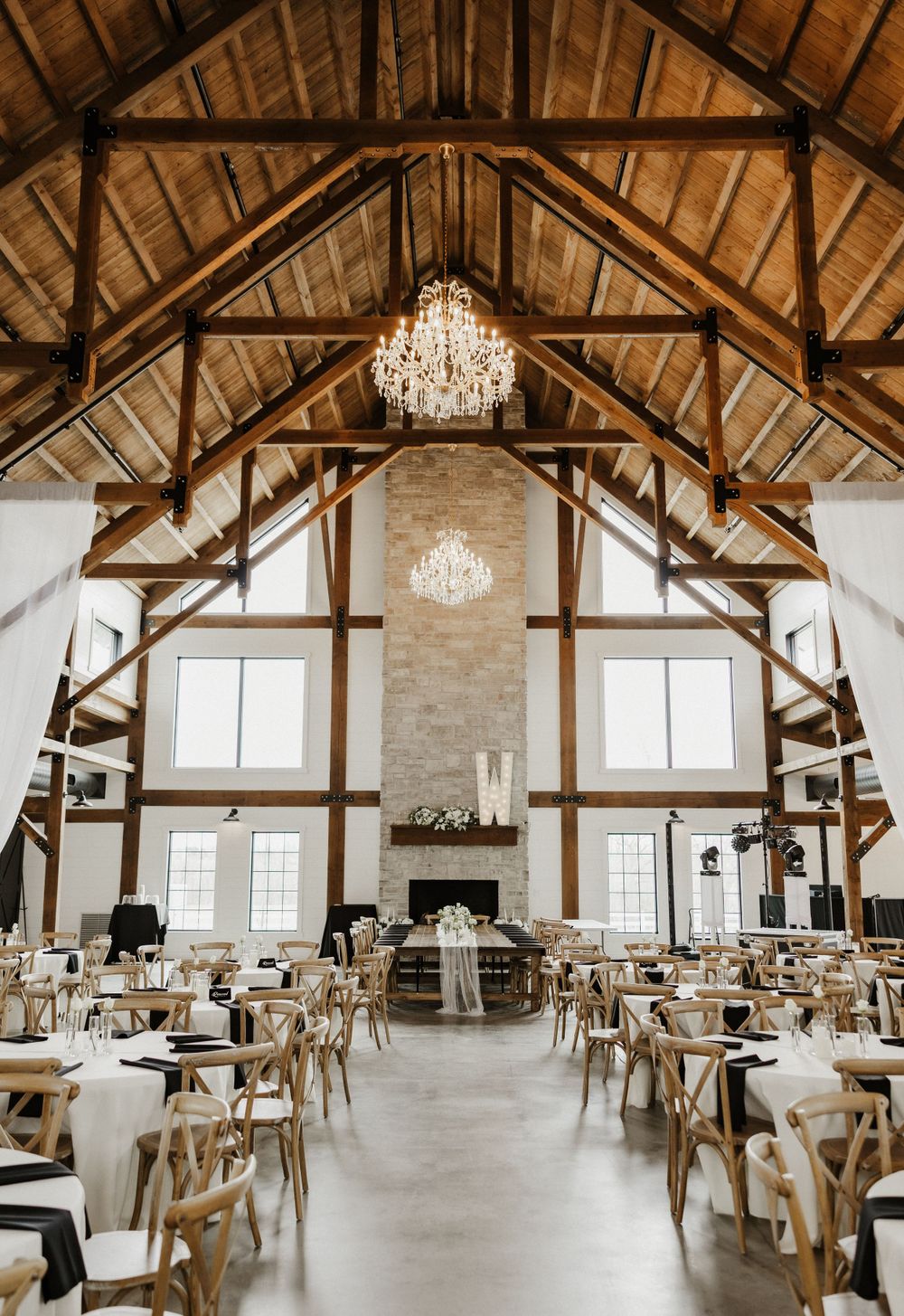 Elegant rustic barn venue with vaulted wooden ceiling, crystal chandeliers, stone fireplace and cross-back chairs for wedding reception.