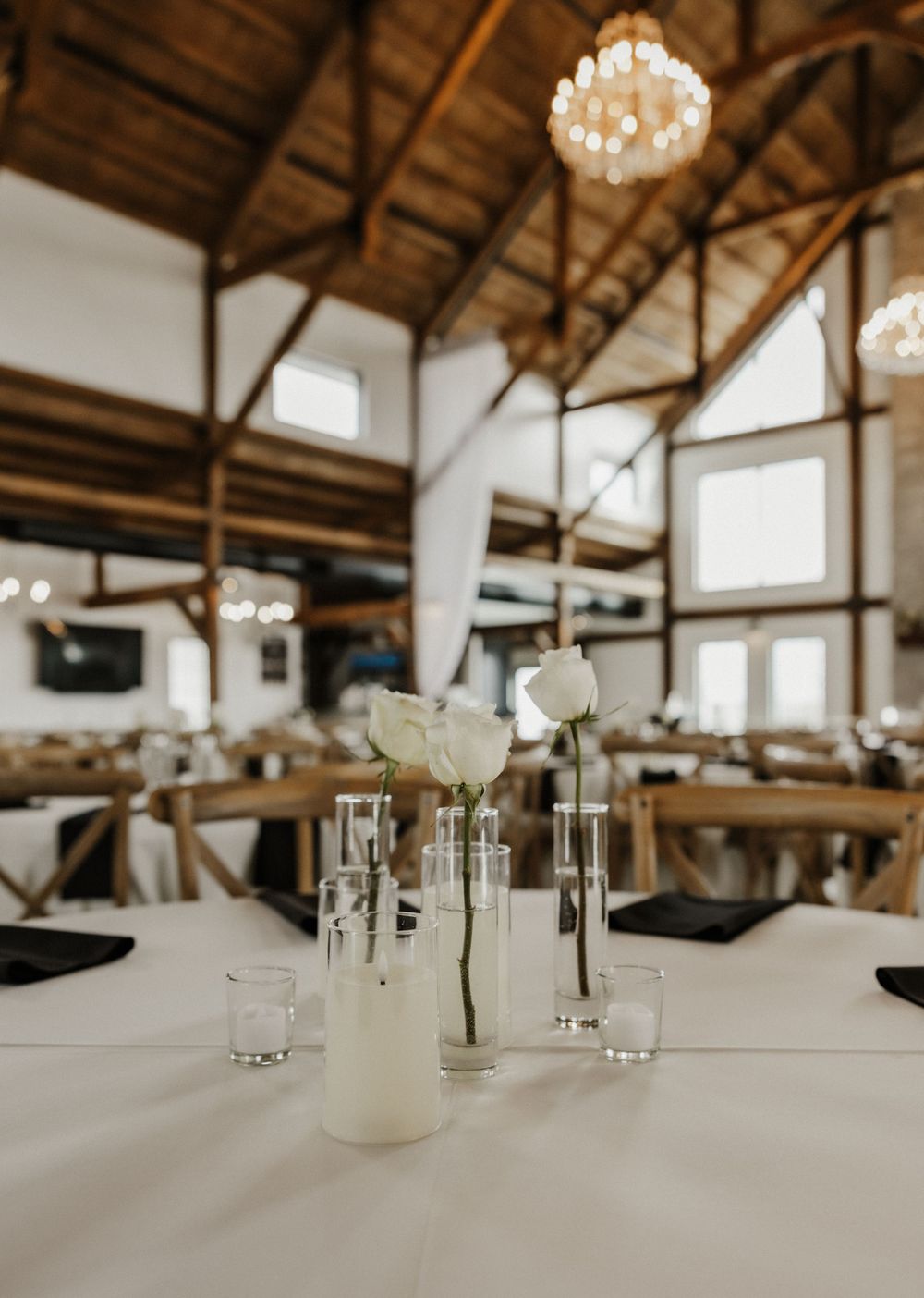 A rustic reception venue with exposed wooden beams, elegant chandeliers and white table settings featuring simple floral centerpieces.