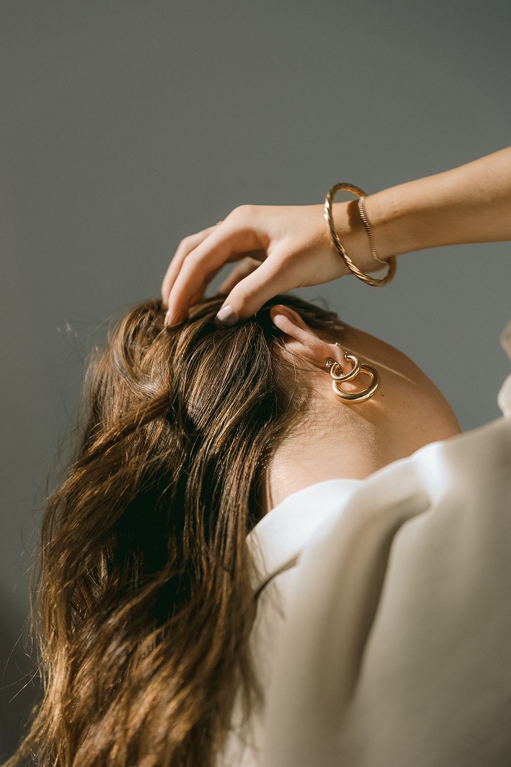A hand wearing a gold bracelet gently touches long flowing brown hair against a gray background.