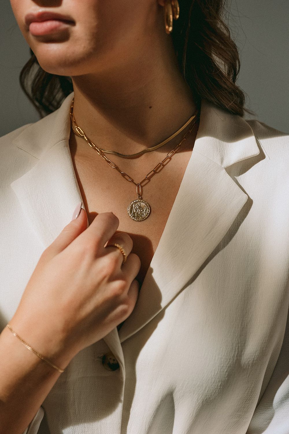 Closeup of layered gold chain necklaces and pendant on a white blazer with delicate rings and jewelry styling.
