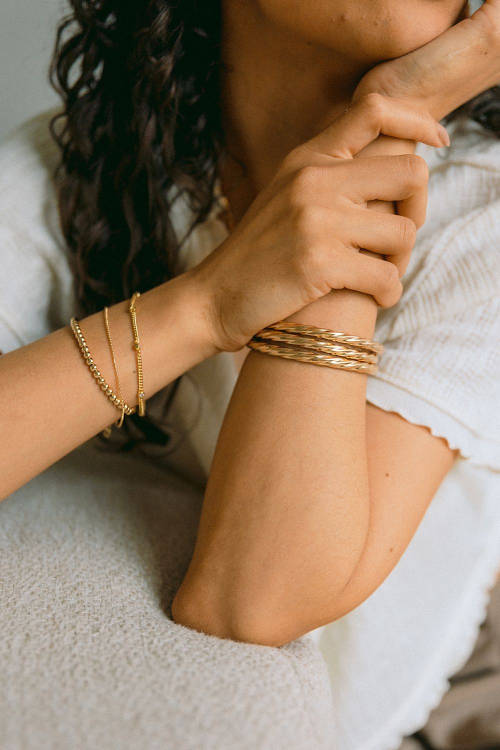 Close-up of hands wearing delicate gold bracelets and bangles on a light-colored textile background.