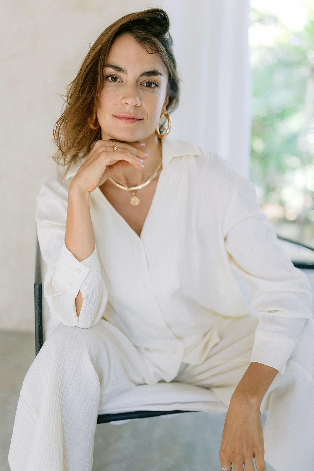 Person in elegant all-white outfit with necklace poses thoughtfully on chair near window in natural light.